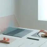 A modern designer workspace features a steel-blue laptop and coral-tinted glass, reflecting liquid glass web design trends inspired by Apple.